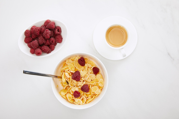 верхний вид чаши кукурузных хлопьев с малиной возле чашки кофе и тарелки со свежими ягодами на поверхности белого мрамора
 - Фото, изображение