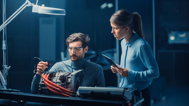 Male and Female Automotive Engineers with a Tablet Computer and Inspection Tools are Having a Conversation While Testing an Electric Engine in a High Tech Laboratory with a Concept Car Chassis. - Fotó, kép