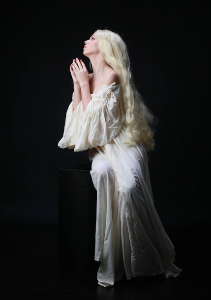 Ghostly portret kobiety z długimi blond włosy ubrany w białą szatę. stwarzające na tle czarnego Studio.  - Zdjęcie, obraz