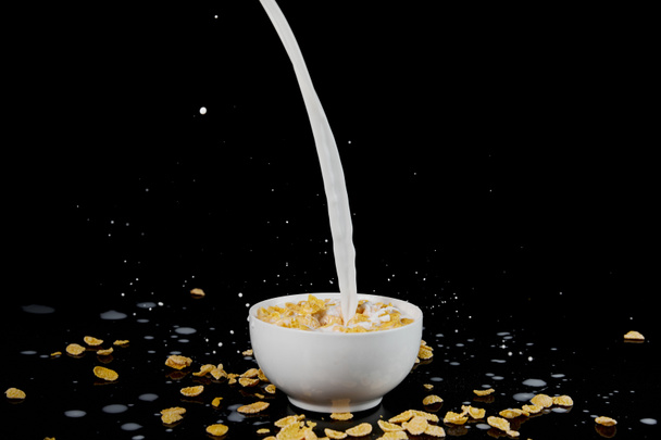 witte kom met cornflakes met melkstroom geïsoleerd op zwart - Foto, afbeelding