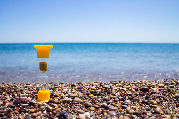 reloj de arena amarillo con arena que se desmorona de pie sobre los guijarros húmedos del mar con el mar y el cielo claro en el fondo
 - Foto, imagen