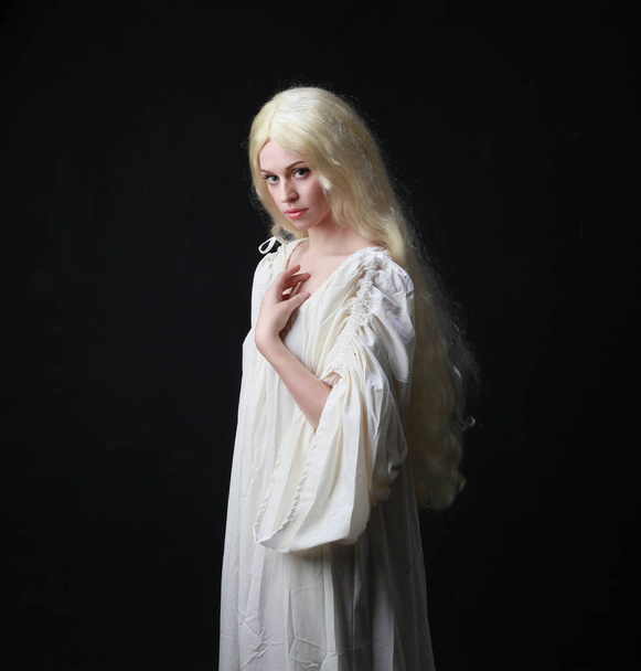 白いローブを着た長いブロンドの髪をした女性の幽霊の肖像画。黒いスタジオの背景に対してポーズ.  - 写真・画像
