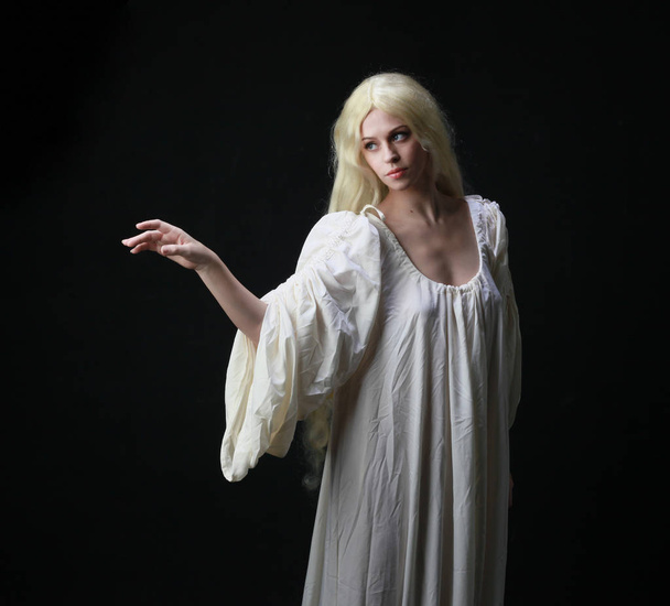 Ένα στοιχειό πορτρέτο μιας γυναίκας με μακριά ξανθά μαλλιά, φορώντας μια άσπρη ρόμπα. ποζάρει σε μαύρο φόντο στούντιο.  - Φωτογραφία, εικόνα