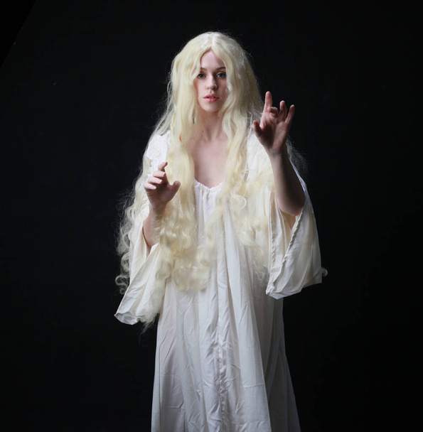Retrato fantasmagórico de uma mulher com longos cabelos loiros vestindo um manto branco. posando contra um fundo estúdio preto
.  - Foto, Imagem