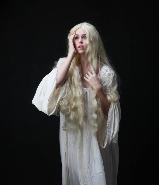 Ghostly portret kobiety z długimi blond włosy ubrany w białą szatę. stwarzające na tle czarnego Studio.  - Zdjęcie, obraz