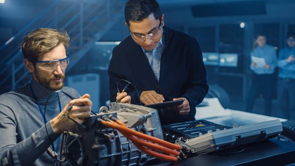 Deux ingénieurs automobiles professionnels dotés d'un ordinateur tablette et d'outils d'inspection discutent lors de l'essai d'un moteur électrique dans un laboratoire de haute technologie doté d'un châssis de voiture concept
. - Photo, image