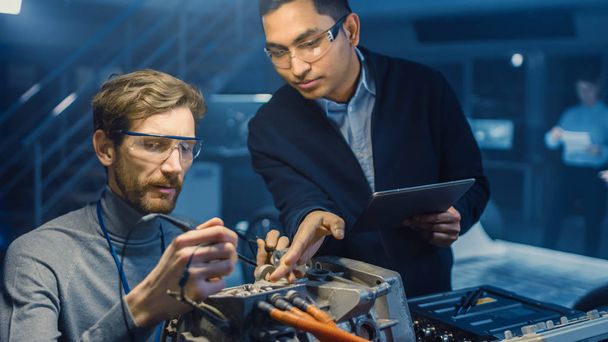 Two Professional Automotive Engineers with a Tablet Computer and Inspection Tools are Having a Conversation While Testing an Electric Engine in a High Tech Laboratory with a Concept Car Chassis. - 写真・画像