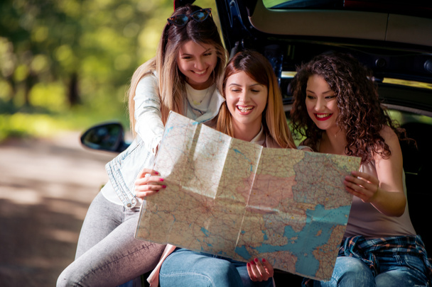 3人の若い女の子が車で休暇に行く。彼らは車のトランクに座って地図を持ってる. - 写真・画像