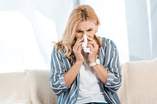 sick woman sitting on sofa and sneezing in napkin  - Φωτογραφία, εικόνα