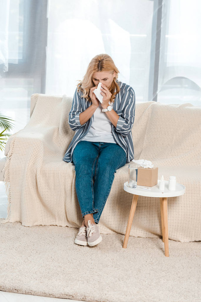 Kranke Frau sitzt auf Sofa und niest in Serviette neben Gewebebox  - Foto, Bild