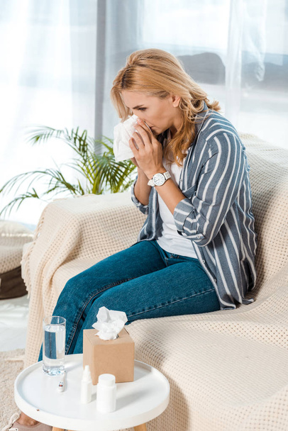 woman with closed eyes sitting on sofa and sneezing while holding tissue  - Photo, Image