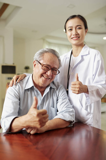 Портрет азиатского пожилого мужчины и молодой женщины-врача, улыбающегося в камеру и показывающего большие пальцы вместе за столом
 - Фото, изображение