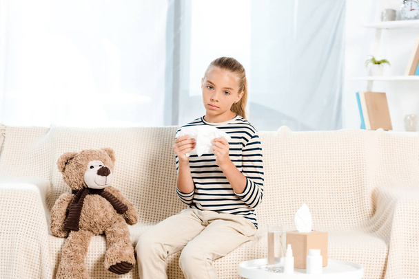 sick kid holding napkin near teddy bear and sitting on sofa  - Фото, зображення