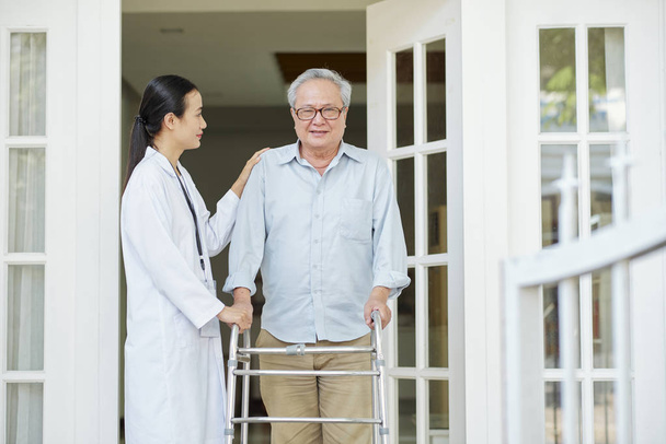 Asian senior man in eyeglasses standing with walker near the entrance of his house and trying to go out with his home caregiver - Photo, Image