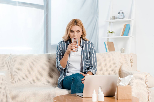 beteg nő iszik vizet, miközben nézi laptop közelében szöveti doboz otthon  - Fotó, kép