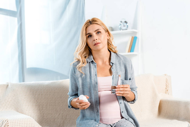 sick pregnant woman sitting on sofa and holding glass of water and bottle with pills - Foto, imagen