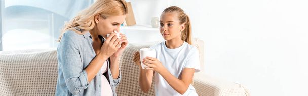 panoramic shot of kid holding cup near mother sneezing in tissue  - Foto, Bild