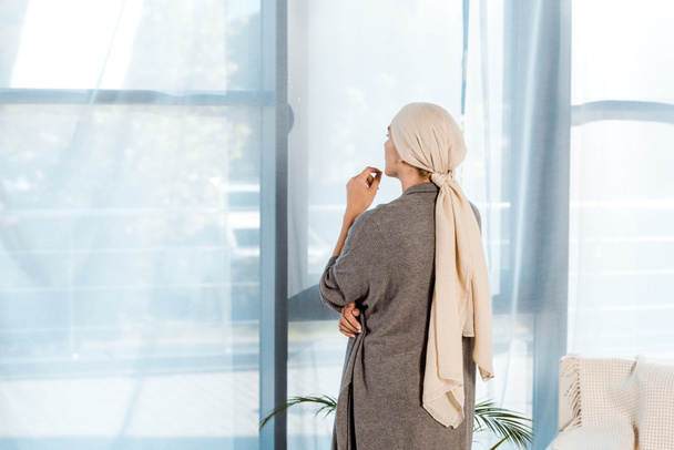 窓の近くのリビングルームに立っている頭のスカーフを持つ病気の女性  - 写真・画像