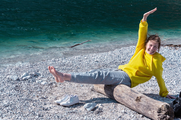 Una chica con una sudadera con capucha amarilla con una sonrisa en la cara se sienta en un tronco junto al lago en las montañas y equilibra
.  - Foto, imagen