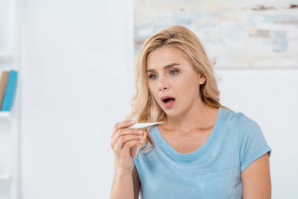 shocked woman holding digital thermometer at home  - Foto, afbeelding