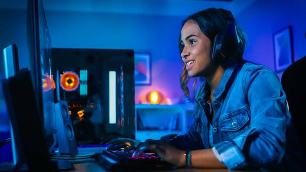 Pretty and Excited Black Gamer Girl in Headphones is Playing First-Person Shooter Online Video Game on Her Computer. Room and PC have Colorful Neon Led Lights. Cozy Evening at Home. - Fotografie, Obrázek
