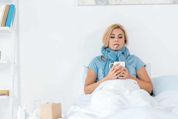 sick woman in blue scarf holding digital thermometer in mouth and cup in hands - Photo, Image