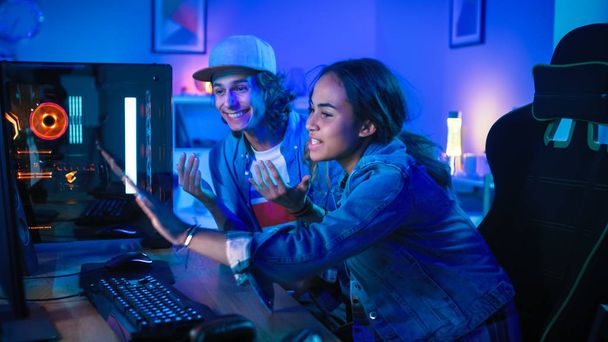 Surprised and Excited Young Man and Girl Bloggers are Watching an Action Video on a Powerful Personal Computer. They are Actively Gesturing. Room and PC have Colorful Neon Led Lights. Cozy Evening at Home. - 写真・画像