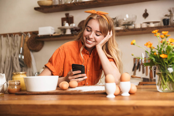 Pleased young blonde girl chef cooking at the kitchen using mobile phone. - Foto, Imagen