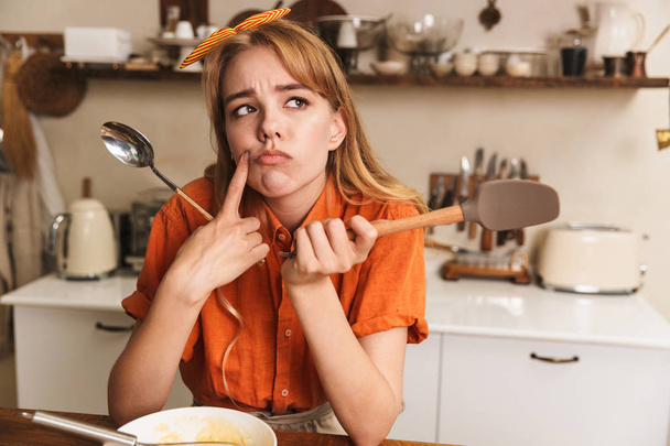 Sad young blonde girl chef cooking at the kitchen thinking. - Foto, Imagen