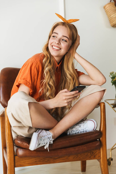 Усміхнена молода блондинка вдома використовує мобільний телефон, сидячи на стільці
. - Фото, зображення