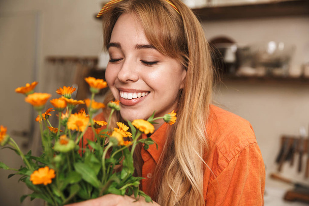Cheerful smiling young blonde girl at the kitchen holding beautiful flowers in hands. - Foto, imagen
