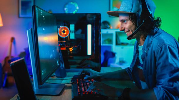 Professional Gamer Playing First-Person Shooter Online Video Game on His Powerful Personal Computer. Room and PC have Colorful Neon Led Lights. Young Man is Wearing a Cap. Cozy Evening at Home. - Foto, Bild