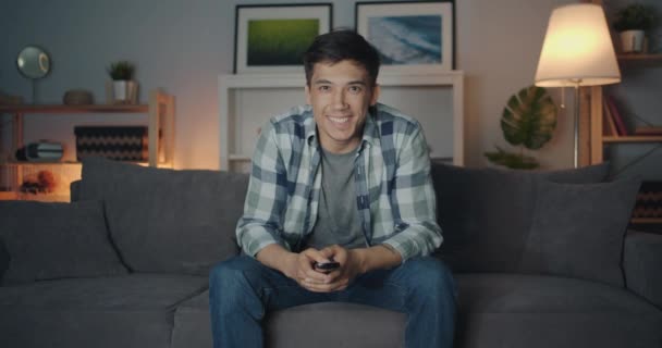 Portrait of mixed race young man watching TV at night laughing having fun - Séquence, vidéo