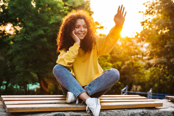 Happy cute young student curly girl sitting on bench outdoors in nature park with beautiful sunlight looking aside waving to friends. - Foto, Imagen