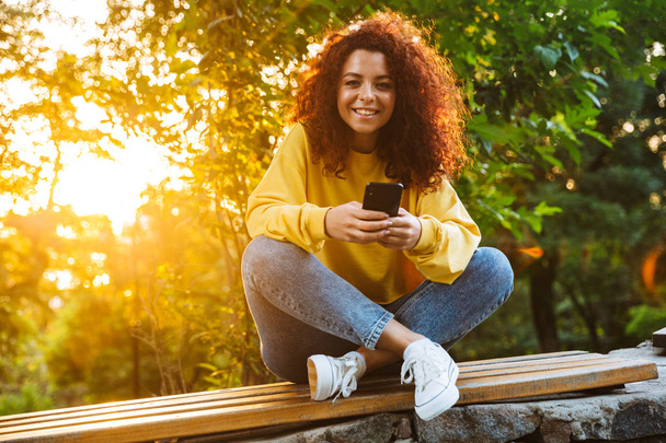 Cheerful smiling cute young student curly girl sitting on bench outdoors in nature park with beautiful sunlight using mobile phone. - Φωτογραφία, εικόνα