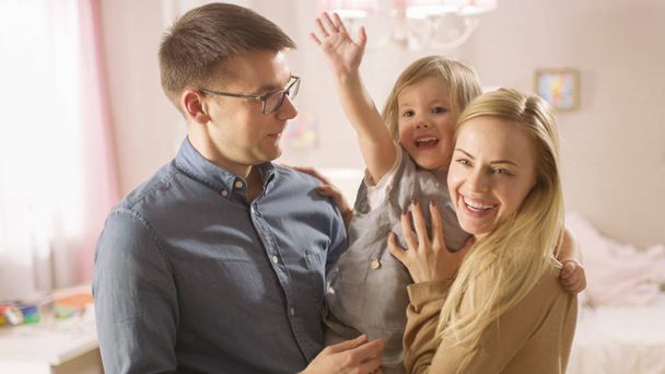 Young Parents Hold Daughter in Their Arms and Hug Her. Daughter Waves to the Camera. They're in a Children's Room which is Pink and full of Toys. - Foto, imagen