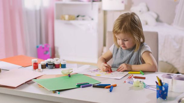 Cute Little Girl Sits at Her Table and Draws with Crayons. Her Room Is Pink, Pretty Drawings Hanging on the Walls, Many Toys Lying Around. - Фото, изображение