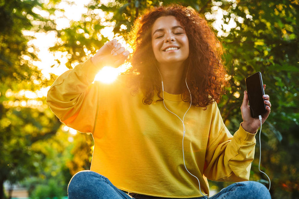 Image of happy young woman listening to music with smartphone and earphones while sitting on bench in green park - Foto, Imagen