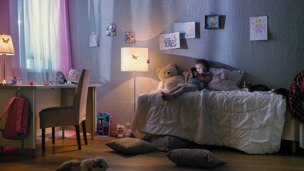 Cute Little Girl Lies on Her Bed and Watches Cartoons on a Smartphone. Her Kitten is Beside Her Playing. Her Floor Lamp is On. - Fotó, kép
