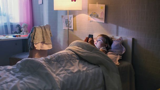 Cute Little Girl Lies in Her Bed and Watches Cartoons on Smartphone. Her Floor Lamp Switched On. - Fotoğraf, Görsel