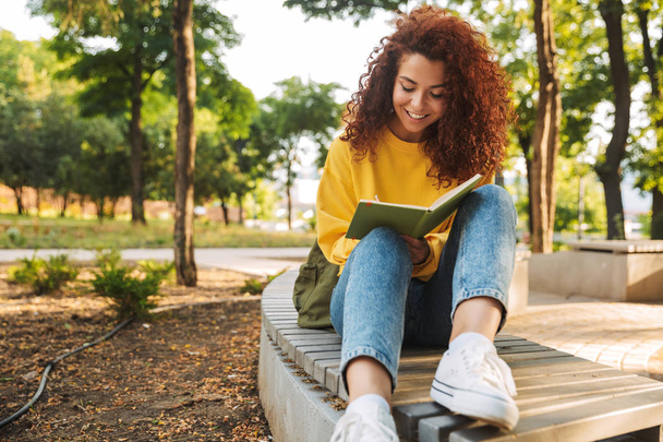 Young beautiful curly student girl sitting outdoors in nature park writing notes in notebook. - Foto, Bild
