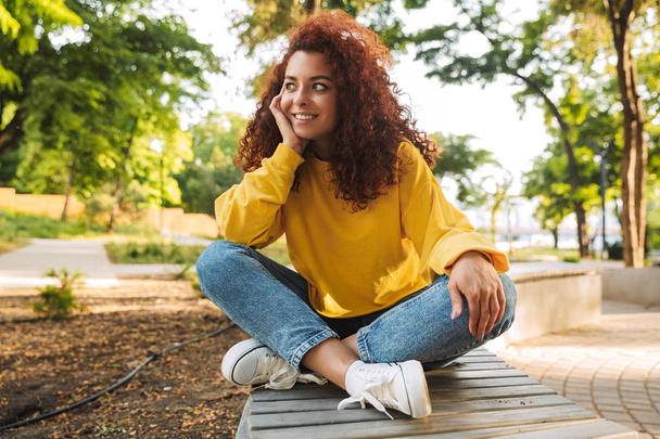 Happy young beautiful curly student girl outdoors in nature park sitting on a bench. - 写真・画像