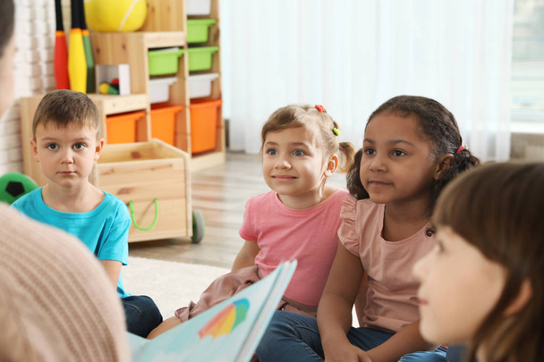 Kindergarten teacher reading book to cute little children indoors - Фото, изображение