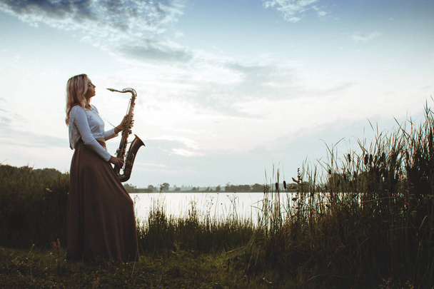 自然を背景にした若い音楽家の肖像、葦の川でサックスを演奏する女性、コンセプト音楽とリラックス - 写真・画像