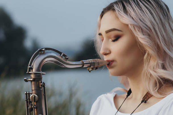 Nehrin kıyısında saksafon çalan genç bir kadın profili, trompete üfleyen bir kadın, doğa arka planı, konsept müzik ve rahatlama. - Fotoğraf, Görsel