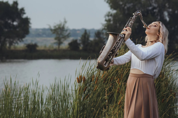 Портрет молодого музиканта на фоні природи, красива жінка грає саксофон на березі річки в очереті, концептуальна музика і розслабтеся
 - Фото, зображення