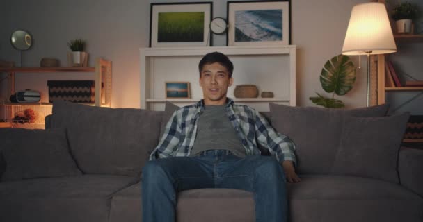 Portrét přitažlivého asijského chlapíka, který sledoval televizi zábavně se smát v tmavé místnosti - Záběry, video