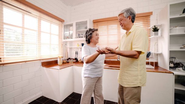 Aziatische ouderen paar dansen samen terwijl ze thuis naar muziek luisteren in de keuken, geniet je van een liefdes moment tijdens het ontspannen thuis. Lifestyle Senior familie ontspannen thuis concept. - Foto, afbeelding