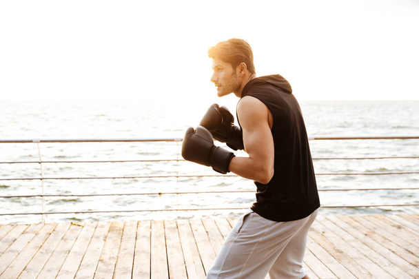 Photo of concentrated man in tracksuit working out in black boxing gloves on wooden pier at seaside - 写真・画像