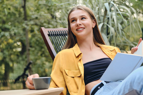 Young pretty brown haired teenage girl in yellow shirt and top happily sitting with book and cup of coffee on wooden deck chair in city park - 写真・画像
